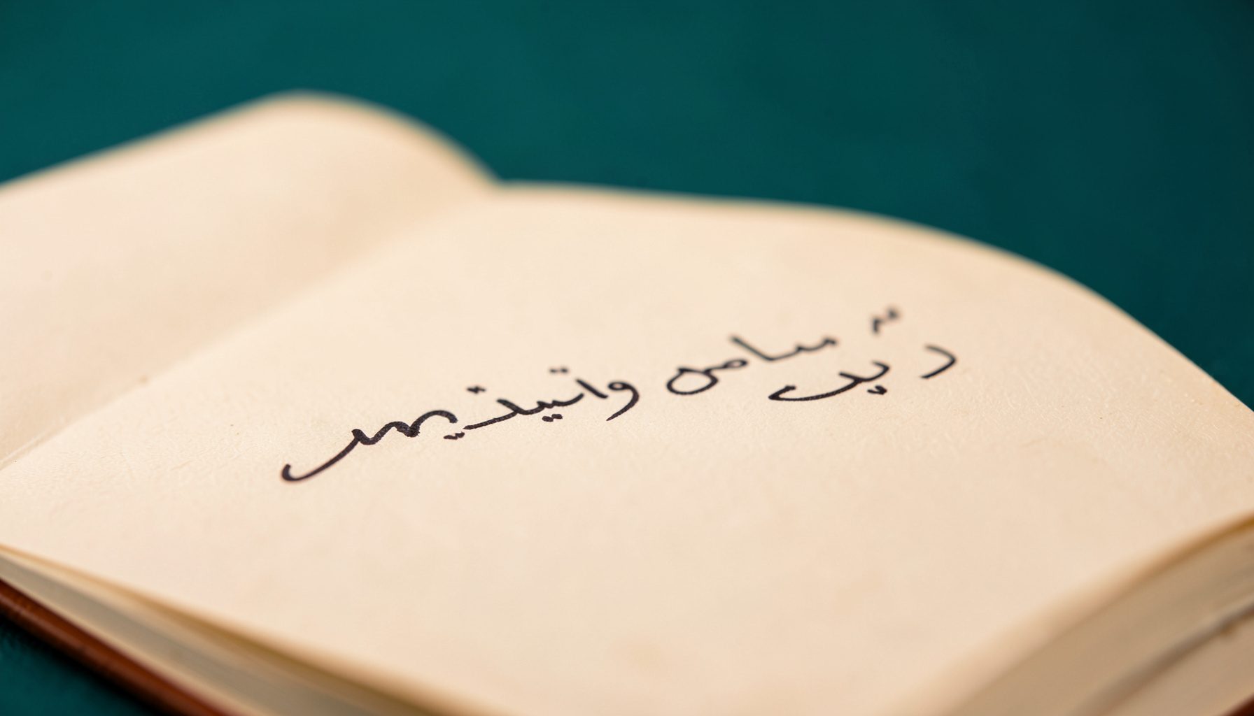 صورة مقربة ليد تكتب بقلم حبر على دفتر مفتوح، الإضاءة دافئة ناعمة والخلفية غير واضحة بلون كريمي